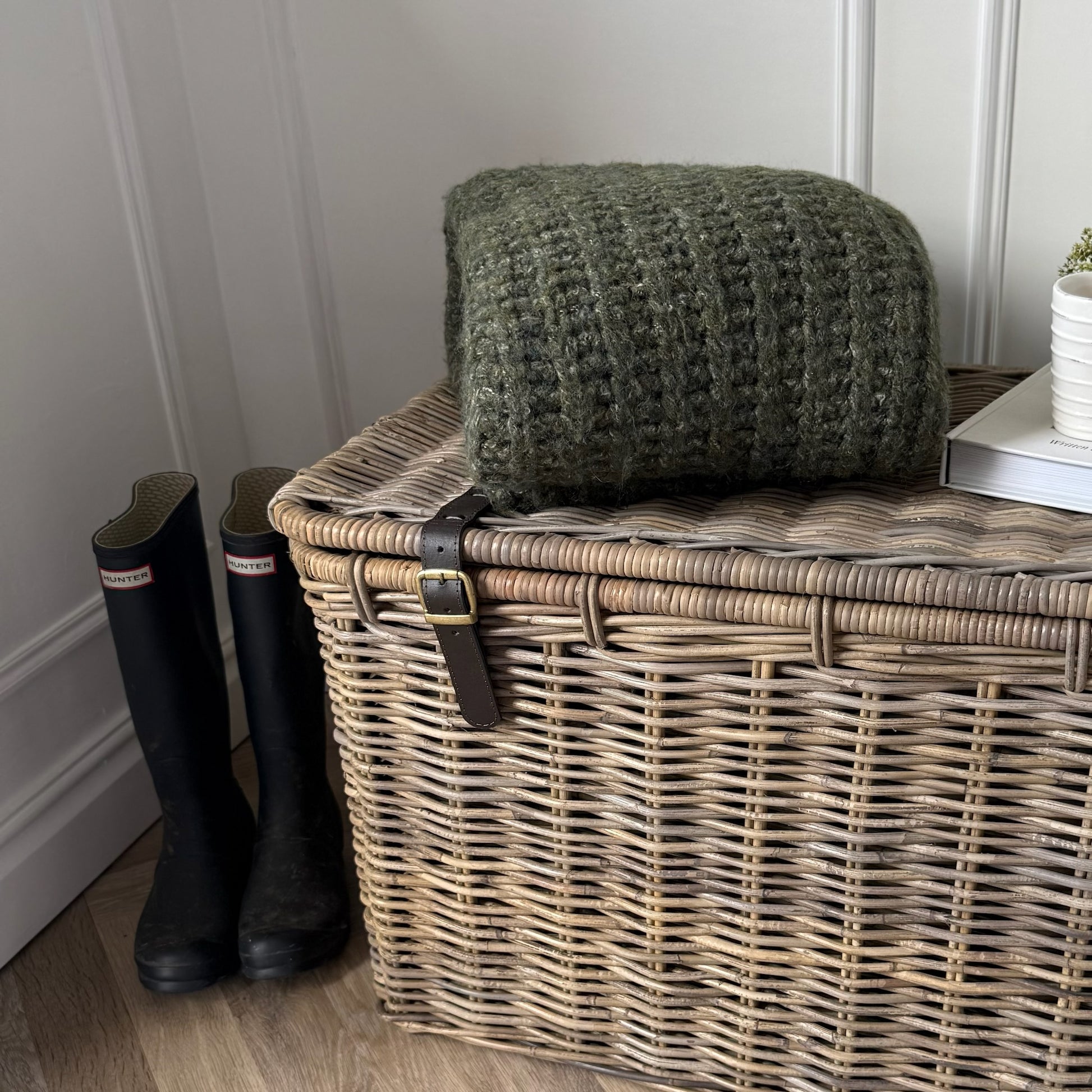 Large wicker trunk with a green chunky blanket and blue hunter wellies on a wooden floor.