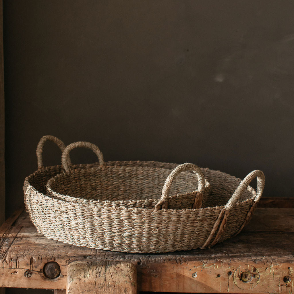 Handwoven natural seagrass basket with plaited handles, styled on a coffee table with books and candles for a warm, rustic look.