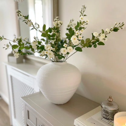 Wild Meadow White Rose Stem with delicate white flowers, soft yellow centres, and realistic green leaves, styled in a vase for timeless home decor. On a console table in a hallway.