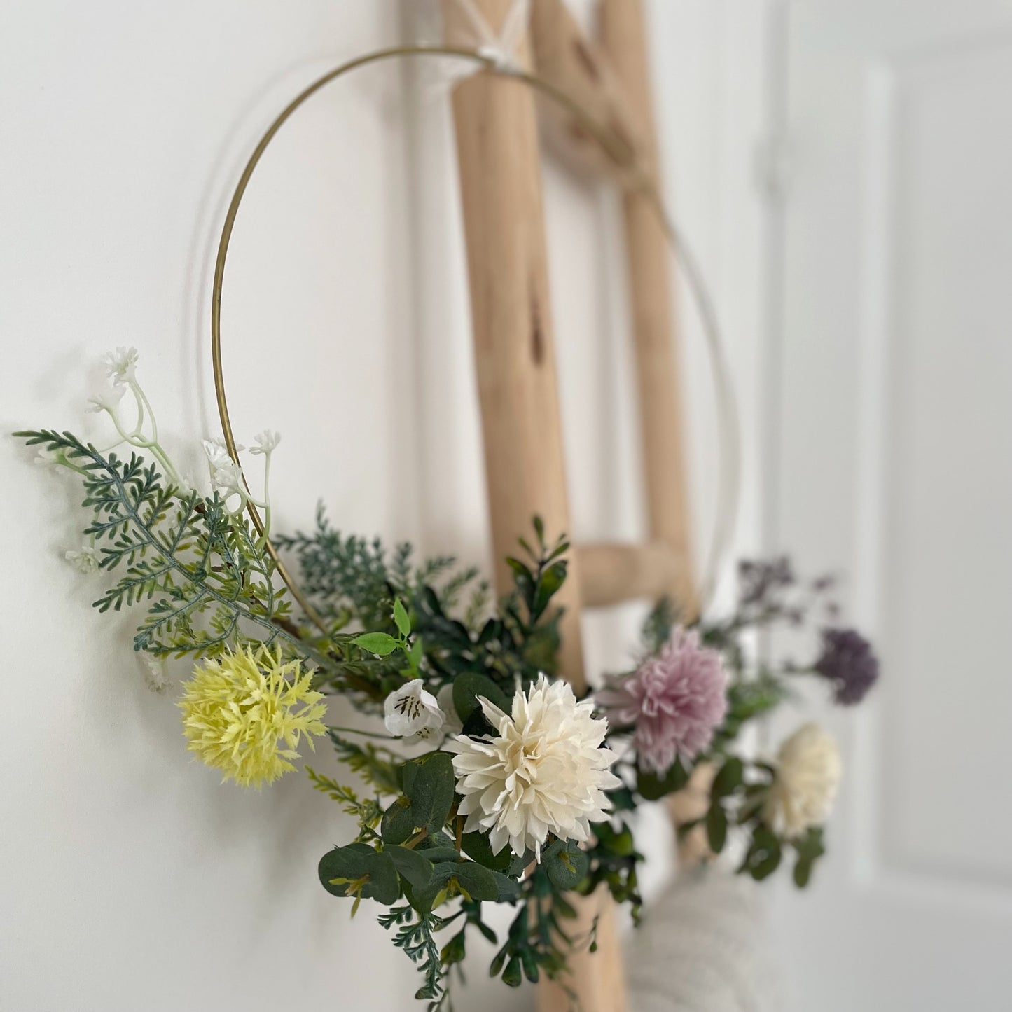 Whimsical hoop wreath decorated with artificial wild flowers and green foliage hanging on a rustic ladder.