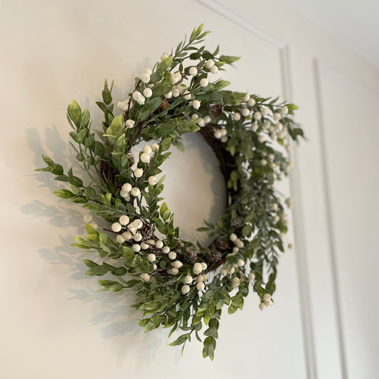 Winter wreath with faux green foliage, white berries and miniature pinecones on a grapevine base, styled on a wall.