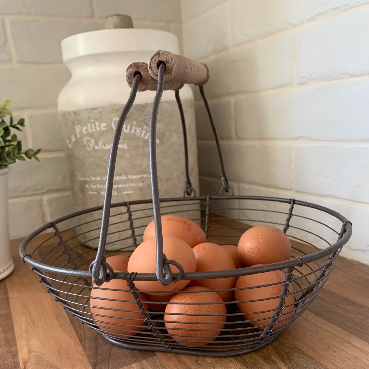 Wire basket with wooden handles holding eggs, styled on a neutral kitchen countertop.