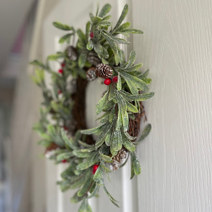 Frosted faux Christmas wreath with green foliage, red berries and fir cones, styled on a white door.