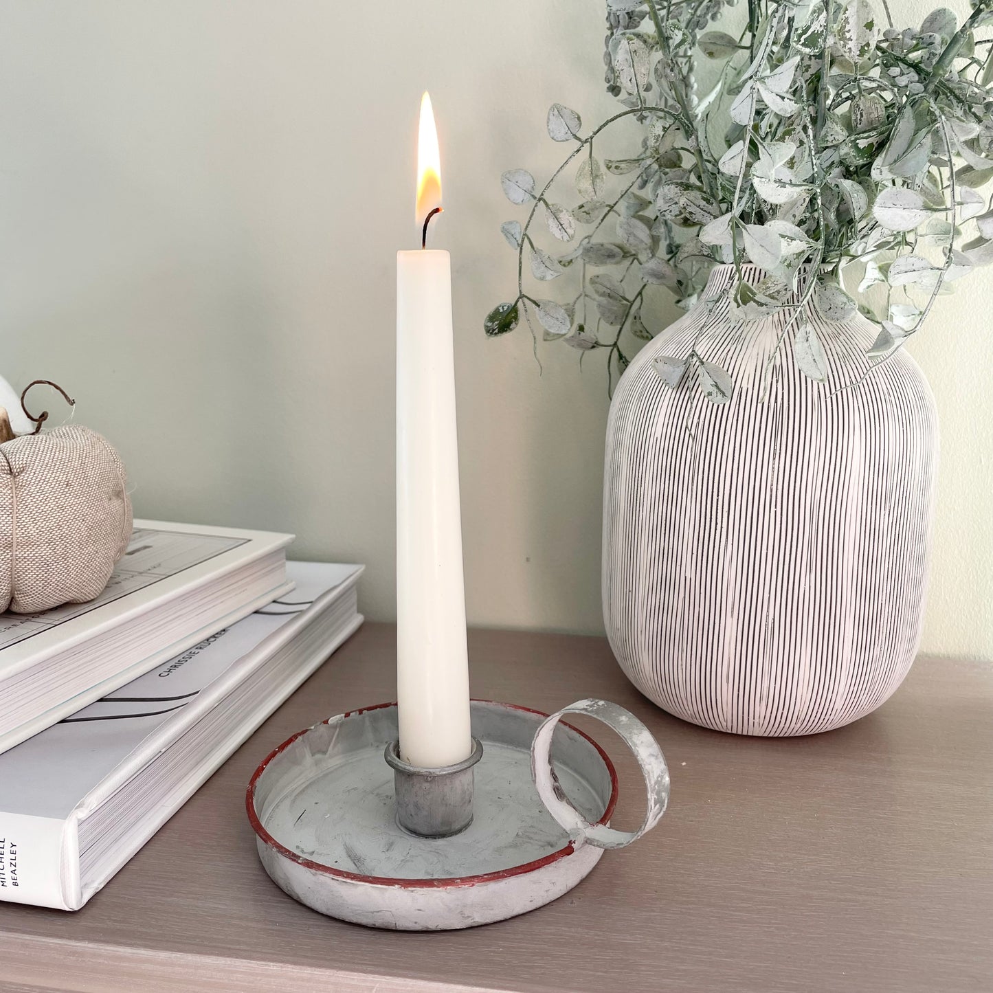 Grey washed wee willie winkie zinc candle holder styled with a taper candle on a wooden console table, with books and a vase of foliage.